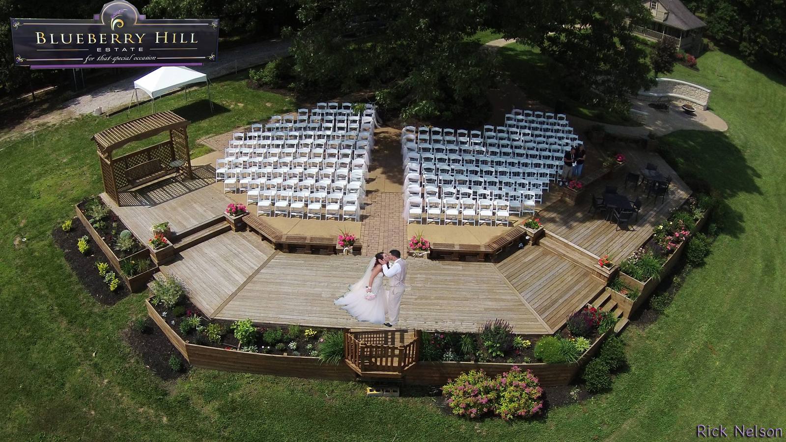 Aerial view of the hilltop ceremony deck — chairs set in rows facing the arbor, the couple on the boards below, and the Blueberry Hill Estate sign at the top of the steps